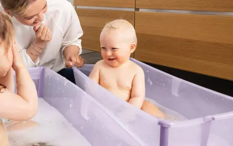 Glad mamma tittar på när ett litet barn njuter av att bada bebis i ett lila badkar med bubblor.