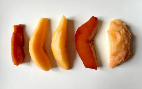 Cinq morceaux de fruit coing cuit, allant du rouge profond à l'orange clair, disposés en ligne sur fond blanc.
