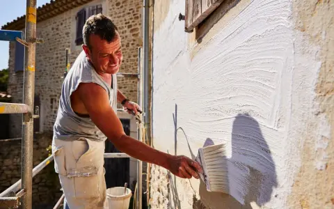 Un homme applique un badigeon blanc sur un mur en pierre avec un large pinceau. Le soleil éclaire la scène, créant des ombres.