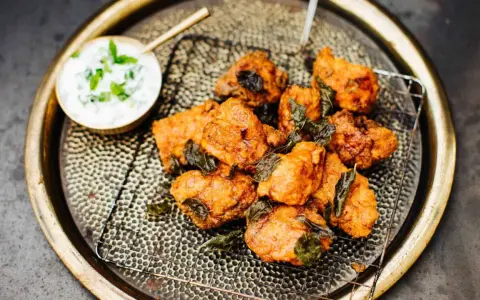 Knusprige chicken pakora auf einem Gitter, garniert mit frischen Kräutern, daneben eine Schale Joghurt-Dip.