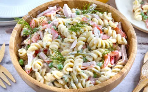 Ein großer Holzteller voller cremigen Nudelsalat mit Schinken, Erbsen und Paprika. Mit Dill garniert, sieht er aus wie der beste Nudelsalat!