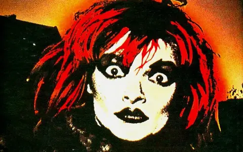 Visage intense d'une femme aux cheveux rouges et maquillage prononcé, évoquant l'esthétique d'un groupe allemand des années 90.