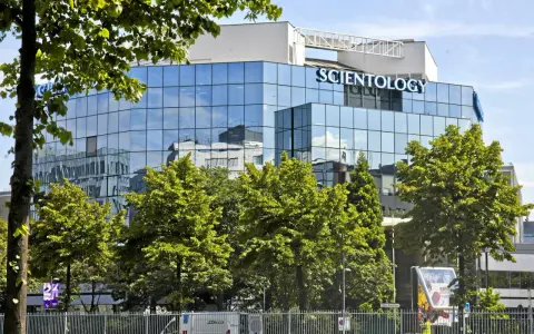 Bâtiment moderne aux façades vitrées reflétant le ciel bleu, avec le nom "SCIENTOLOGY" inscrit en lettres bleues lumineuses. Des arbres verdoyants entourent la structure.