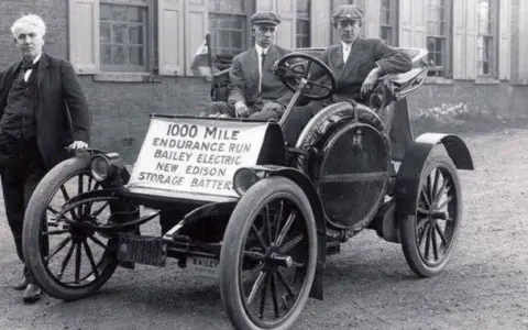 Un hombre junto a un coche eléctrico Bailey de 1907, parte de la historia del coche eléctrico, con un cartel que anuncia una carrera de resistencia de 1000 millas.