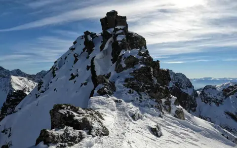 Śnieżny szlak prowadzi na skalisty szczyt, który przypomina zadni granat. W tle widać pasmo górskie.