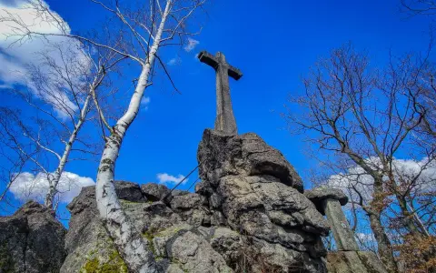 Kamienny krzyż góruje nad skalistym szczytem. Wokół rosną nagie drzewa, a niebo jest błękitne z białymi chmurami.