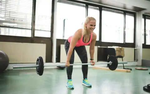 En kvinna lyfter skivstång i ett gym. Hon ler och ser fokuserad ut, en bra start på hur blir man stark.