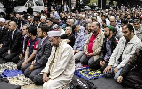 Männer in traditioneller Kleidung knien auf Gebetsteppichen. Ein Mann in einem weißen Gewand und Turban leitet das Gebet, ein Zeichen der berberischen Religion.