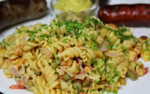 Ein cremiger nudelsalat mit mayo, garniert mit Schnittlauch. Daneben liegen zwei gebratene Würstchen und ein Schälchen Senf.