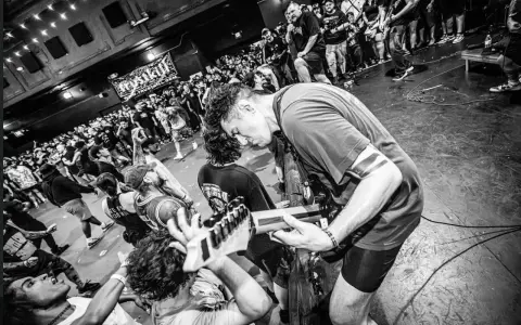 Energetyczny koncert w klubie. Czarno białe zdj. z tłumem szalejącym pod sceną, gdzie gitarzysta pochyla się nad instrumentem.