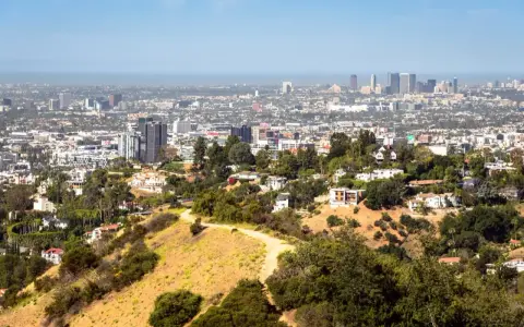 Widok na Los Angeles, miasto śmierci, z wzgórz porośniętych suchą trawą i drzewami. W oddali widać drapacze chmur.