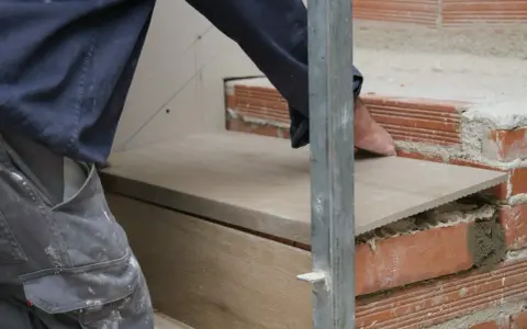 Układanie drewnopodobnych płytek na schodach. Rzemieślnik precyzyjnie dopasowuje cokół na schodach z płytek.