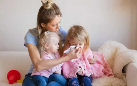 Mama wyciera nos chorej córce, podczas gdy młodsza siostra patrzy. Czy szczepienie mogłoby zapobiec katarowi?