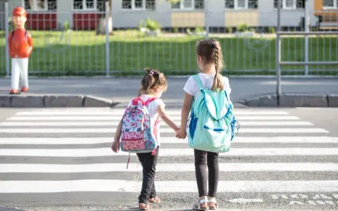 Dwie dziewczynki z plecakami idą za rękę przez pasy. Od ilu lat dziecko może samo chodzić po mieście?