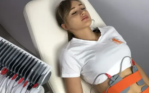 Kobieta leży na kozetce, poddana zabiegowi elektrostymulacji brzucha. Elektrody przymocowane są do jej talii.