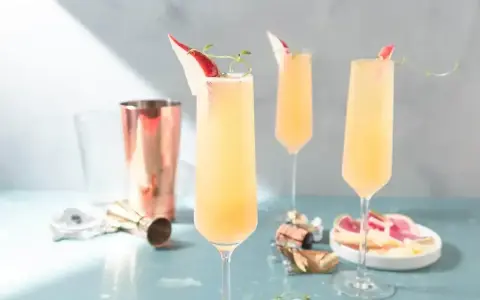 Tre eleganta glas med en ljusgul drink med gin och prosecco, garnerade med en tunn skiva röd frukt och en kvist.