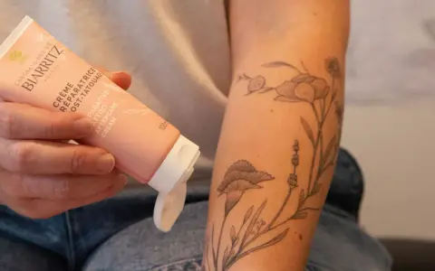 Une main tient un tube de crème réparatrice post-tatouage, prêt à être appliqué sur un bras orné d'un tatouage floral délicat.