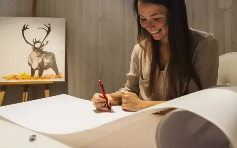 En kvinna ler medan hon ritar på ett stort papper. I bakgrunden syns en oljemålning av en ren. Hon deltar i en oljemålning kurs.