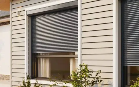 Volet roulant gris fermé sur une fenêtre de maison avec bardage beige.