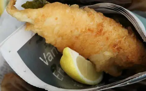 Krispigt friterad fisk med citronklyfta, inslagen i tidningspapper. En klassisk fish and chips-upplevelse.