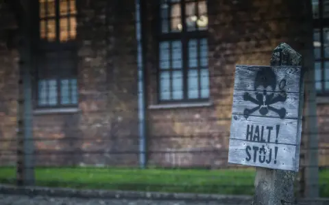 Tablica z symbolem czaszki i piszczeli oraz napisem "HALT! STÓJ!" na tle ceglanego budynku i drutu kolczastego, przypominająca o grozie obozów koncentracyjnych.