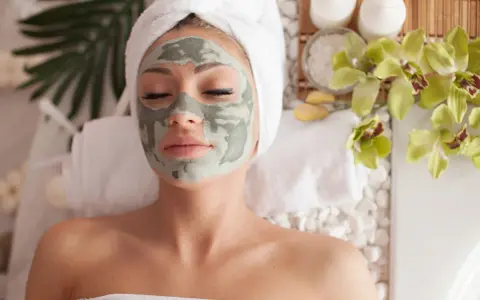 Femme détendue avec un masque fait maison, enveloppée dans une serviette, entourée de fleurs d'orchidée et de bougies.