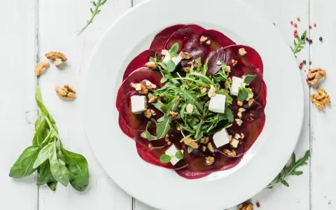 Eleganckie carpaccio z buraka z rukolą, orzechami włoskimi i kostkami sera feta na białym talerzu.