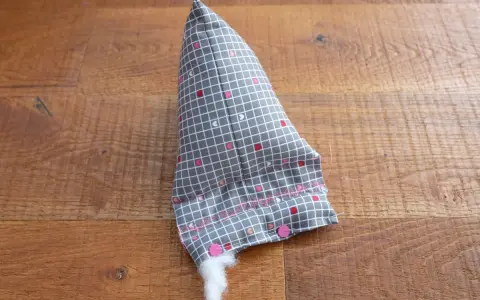 Un berlingot tissu gris à petits cœurs roses et blancs, en cours de rembourrage, est posé sur un parquet en bois.