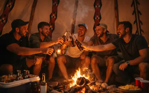 Pięciu uśmiechniętych mężczyzn wznosi toasty butelkami piwa przy ognisku. Niezapomniany wieczór kawalerski w górach.