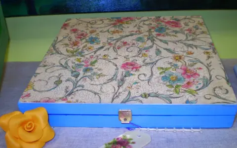 Caja azul con tapa decorada con papel decoupage floral. Un detalle de rosa amarilla y etiqueta floral completan la escena.
