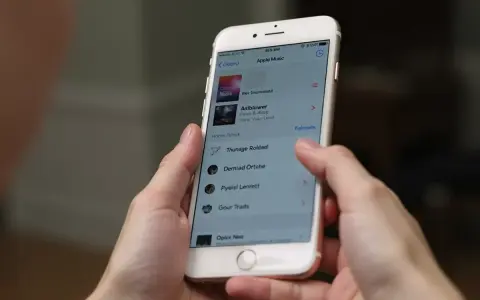 Ręce trzymają iPhone'a z otwartą aplikacją Apple Music. Widoczna lista utworów, która może pomóc w nauce, jak pobrać muzykę na iPhone'a.