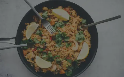 En stor panna med jollof rice, räkor, citronklyftor och färsk persilja. Tre bestick ligger redo att avnjutas.