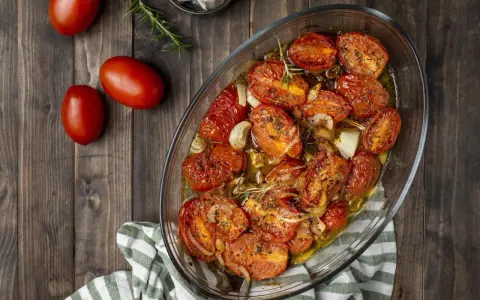 Pomodori gratinati al forno, dorati e succosi, con cipolle e rosmarino in una teglia di vetro su un tavolo di legno.