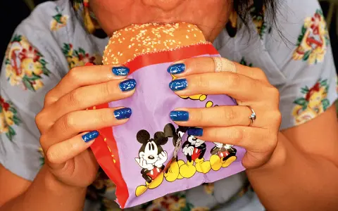 Mani con smalto blu intenso tengono un hamburger in un involucro con Topolino, in uno stile tipico di Martin Parr.