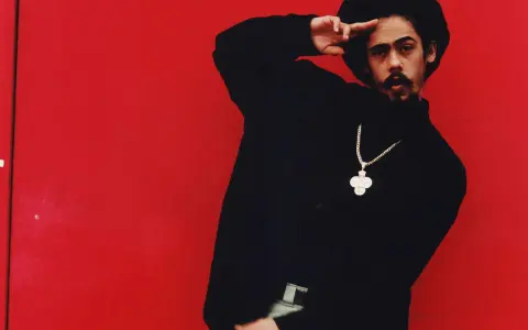 Mann mit Afro und Schnurrbart, schwarzer Rollkragenpullover, goldene Kette mit Kreuzanhänger, vor rotem Hintergrund. Er salutiert. "I shot the sheriff" könnte man sagen.