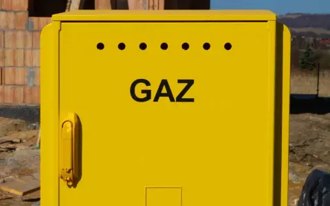 Żółta szafka gazowa z napisem "GAZ" i otworami wentylacyjnymi. Obok budowa domu, w tle krajobraz. Przyłącze gazowe krok po kroku.