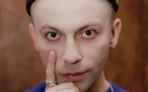 Zbliżenie na twarz Arka Kłusowskiego w czarnej czapce, z palcem przy policzku.