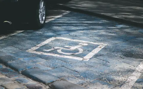 Niebieskie miejsce parkingowe dla niepełnosprawnych pod blokiem, z symbolem wózka inwalidzkiego. Obok widoczna część czarnego samochodu.