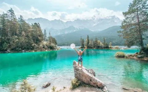 Frau mit ausgebreiteten Armen und Hut am türkisfarbenen Eibsee in Bayern, im Hintergrund Berge und Wald.