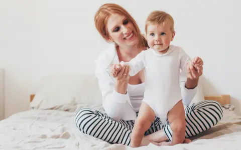 Uśmiechnięta mama pomaga swojemu 8 miesięcznemu dziecku w pierwszych krokach na łóżku.