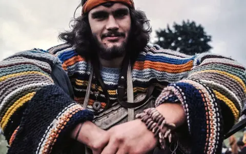 Młody mężczyzna w kolorowym swetrze w paski i opasce na głowie, uśmiecha się. Wygląda jak prawdziwy hippie.