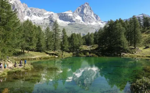 Un gruppo di escursionisti ammira il riflesso del Cervino nel lago blu di Cervinia, circondato da pini e montagne innevate.