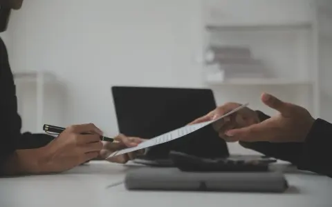 Dwie osoby omawiają dokument, jedna wskazuje piórem na klauzulę CV. Obok leży laptop i kalkulator.