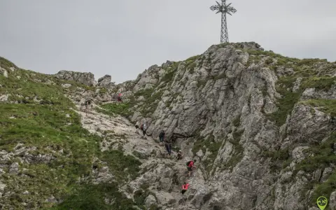 Grupa turystów wspina się po skalistym zboczu w kierunku krzyża na szczycie. Trudno określić, ile idzie się na Giewont, ale widok jest imponujący.