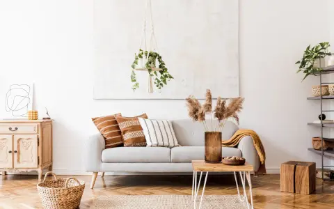Un salón moderno con un sofá gris, cojines étnicos y una manta mostaza. La decoración se completa con una planta colgante y un jarrón con hierbas secas.