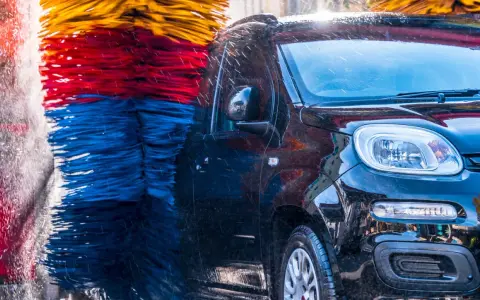 Czarna, błyszcząca karoseria samochodu w myjni automatycznej, otoczona kolorowymi szczotkami. Czy myjnia automatyczna niszczy lakier? Woda spływa po aucie.