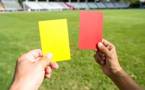Sędzia pokazuje żółtą kartkę, która oznacza ostrzeżenie w piłce nożnej. Obok czerwona kartka.