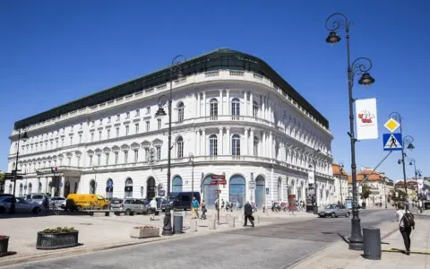 Elegancki, biały budynek w centrum Warszawy, być może najdroższy hotel w Warszawie. Słoneczny dzień, ruch uliczny.