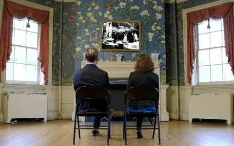 Coppia seduta di spalle, guarda un quadro con una scena di giardino, "Roundhay Garden Scene", su una parete fiorita.