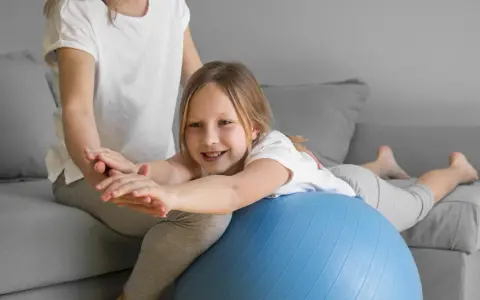 Dziecko z obniżonym napięciem mięśniowym ćwiczy mowę, leżąc na piłce rehabilitacyjnej pod opieką terapeuty.